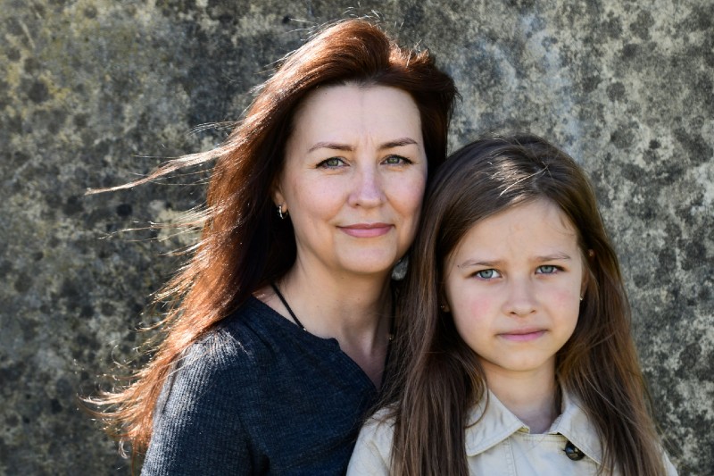 Mayya Simonova. Ella and Dasha, Melbourne, September 2022. Ella fled Cherkasskaya-Lozova village near Kharkiv with her daughter Dasha, soon after bombing began. She left behind her husband and 18-year-old son. A colour portrait of a mother and her eight-year old daughter. The mother is in the centre of the photograph. She has green eyes and long dark auburn hair, and gazes at the camera with her lips closed. The light catches on her hair blowing in the wind. Her daughter sits to her right. She has long medium brown hair and blue eyes. The background is mottled grey, and looks like an old stone wall.