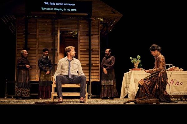 Image shows a stage scene, with a man in a suit without a jacket sitting on the stoop of a house scene to the left and a woman  in a long dress sitting in a chair to the right of screen.