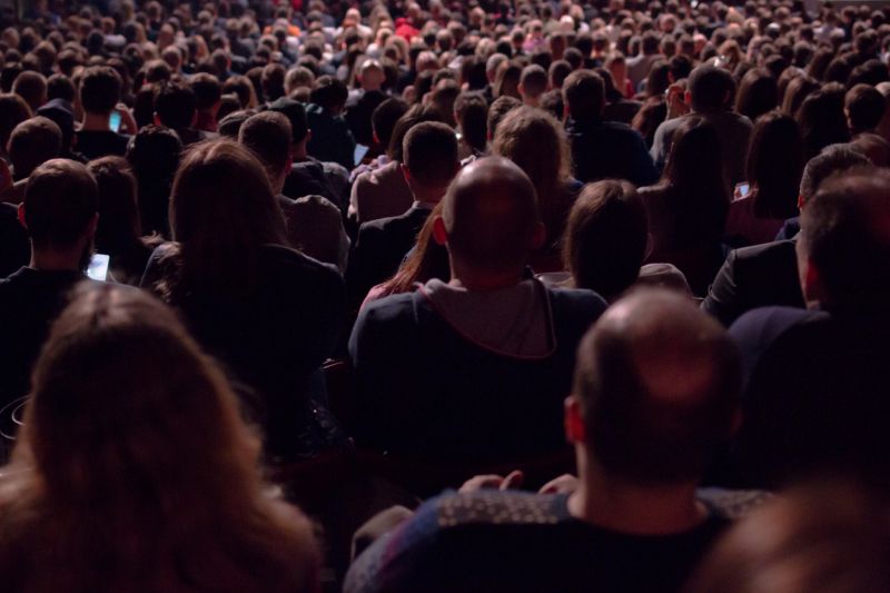 In the dark hall there is a view from the back of a crowd of hundreds of people sitting and watching the screen in a movie theater or performing on stage. Concept for articles