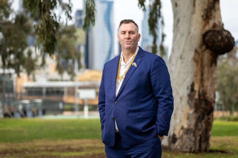 Image of Travis in navy suit standing in front of a tree and park