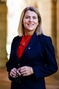 Andrea Carson stands in the Old Quad cloisters wearing a navy suit and red shirt 