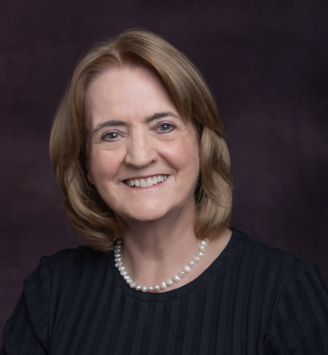 image of smiling woman with a pearl necklace and blonde hair infront of purple backdrop