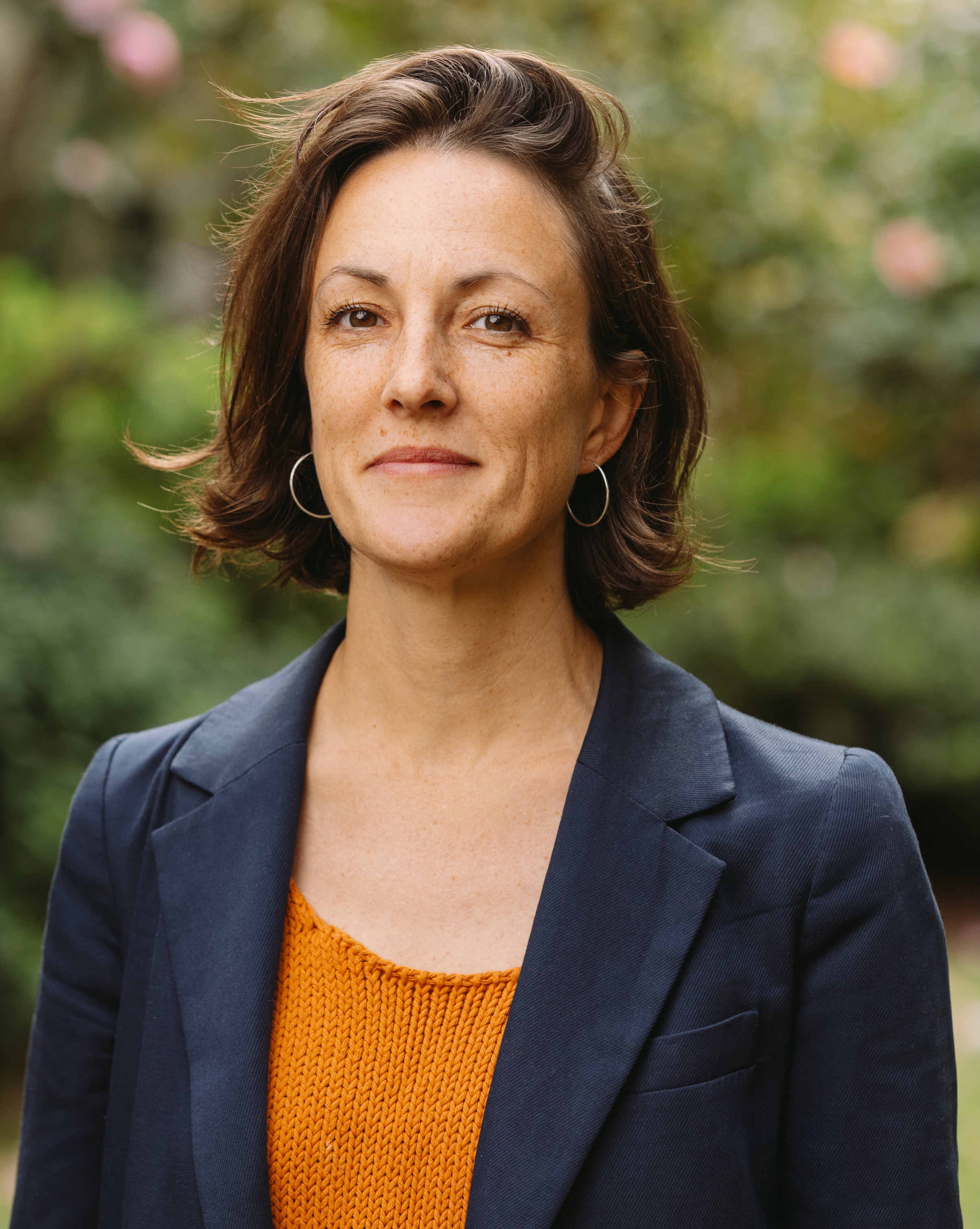 Image shows a wmoman with brown hair above the shoulders, smiling close mouthed, wearing an orange top under a blazer and small hoop earrings, in front of a garden background.