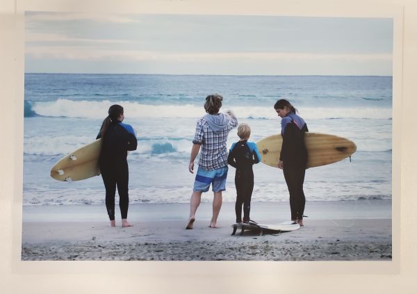 adult standing on oceans edge with 3 children in wetsuits and surfboards
