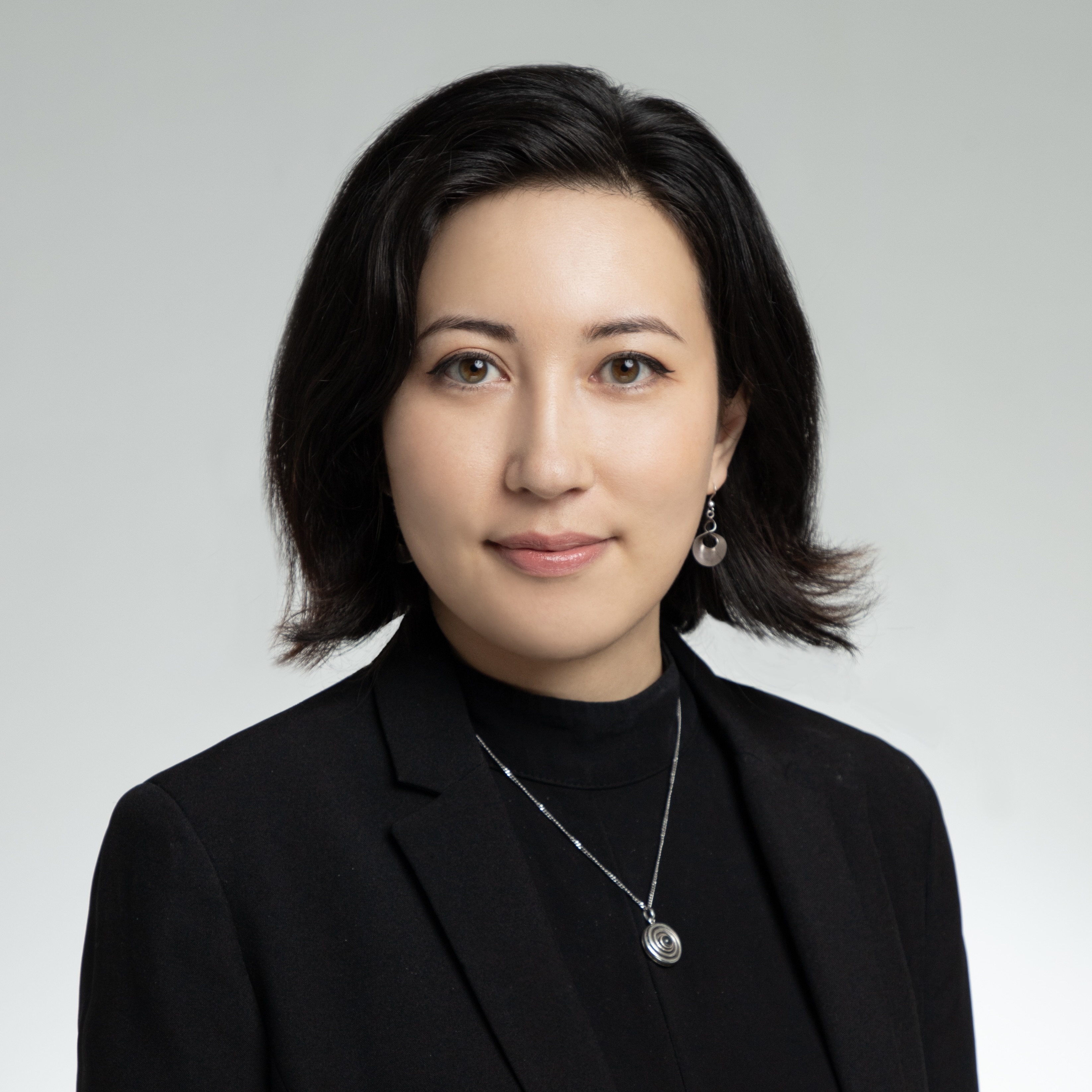 Image shows a headshot of a woman with black hair above the shoulders, wearing black with a silver necklace and earrings.