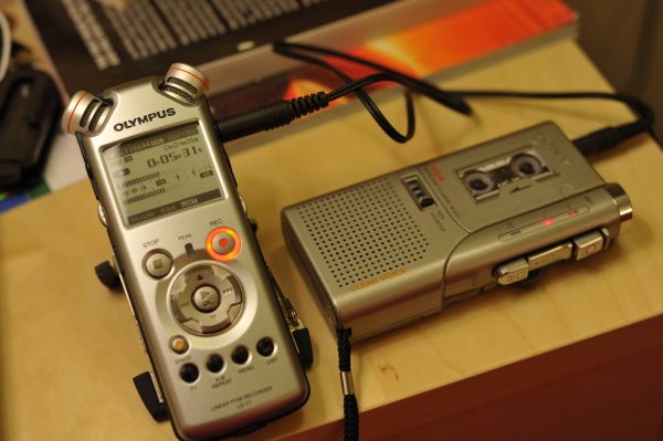 A silver Olympus dictaphone and recording device are displayed placed on a table beside some blurred books. 