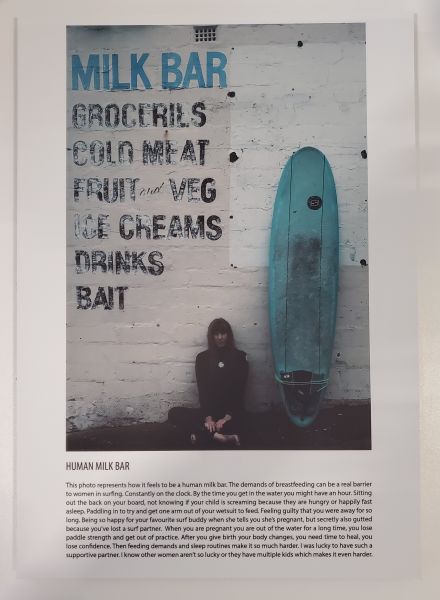 woman sat in wetsuit against a wallk that reads milk bar with blue surfboard next to her