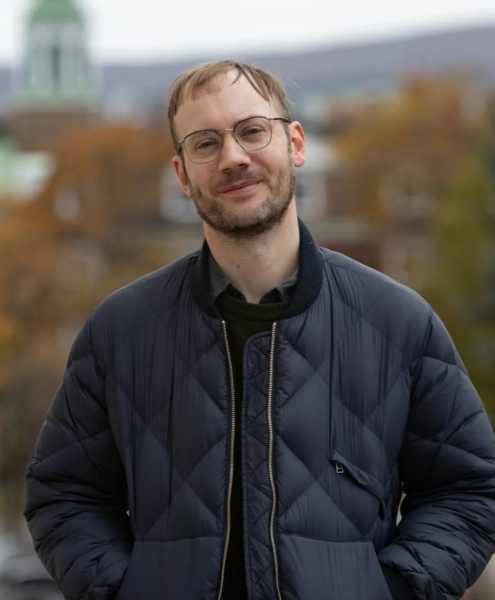 image of white man with glasses, wearing a navy blue jacket in front of blurred greenery and buildings
