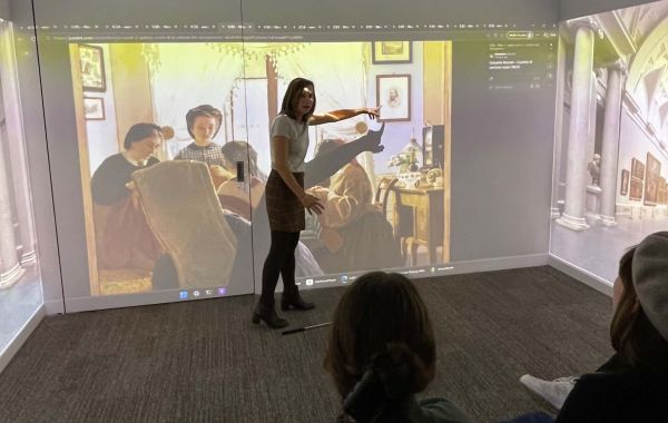 Presenter gestures toward large interactive display showing classical painting with audience members seated in foreground.