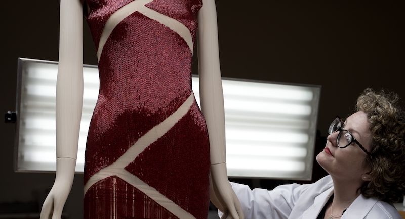 A female conservator with fair skin and short curly hair adjusts a red dress on a mannequin.