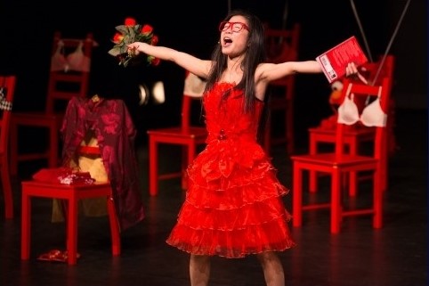 Woman singing in red dress and glasses with arms wide open on a stage wtih a background of red chairs