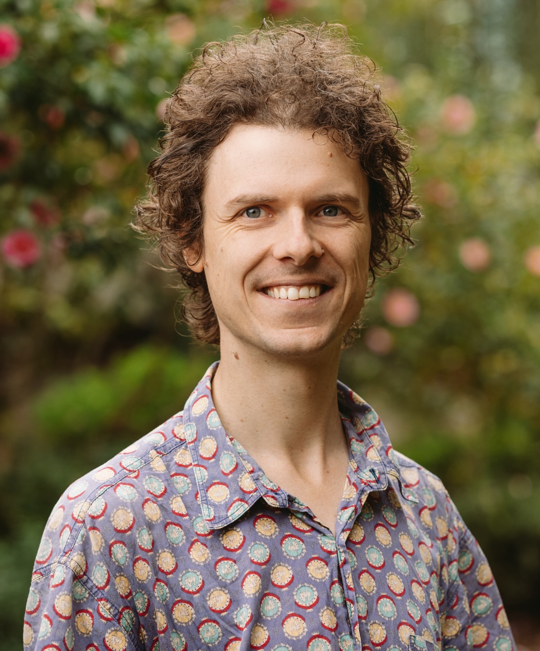 image of smiling man with curly brown hair infront of greenery