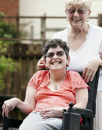 A woman wearing sunglasses, a pink shirt and grey pants sitting in a wheelchair with a woman in a white shirt standing behind her. Both women are laughing.