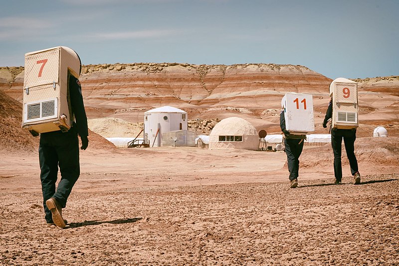 A photo of three researchers in spacesuits approaching the Mars Desert Research station. The station is set in a sandy desert landscaoe.