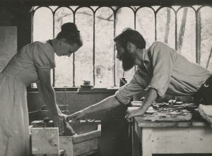 An old photo of a man and woman looking down at a pottery wheel with clay.