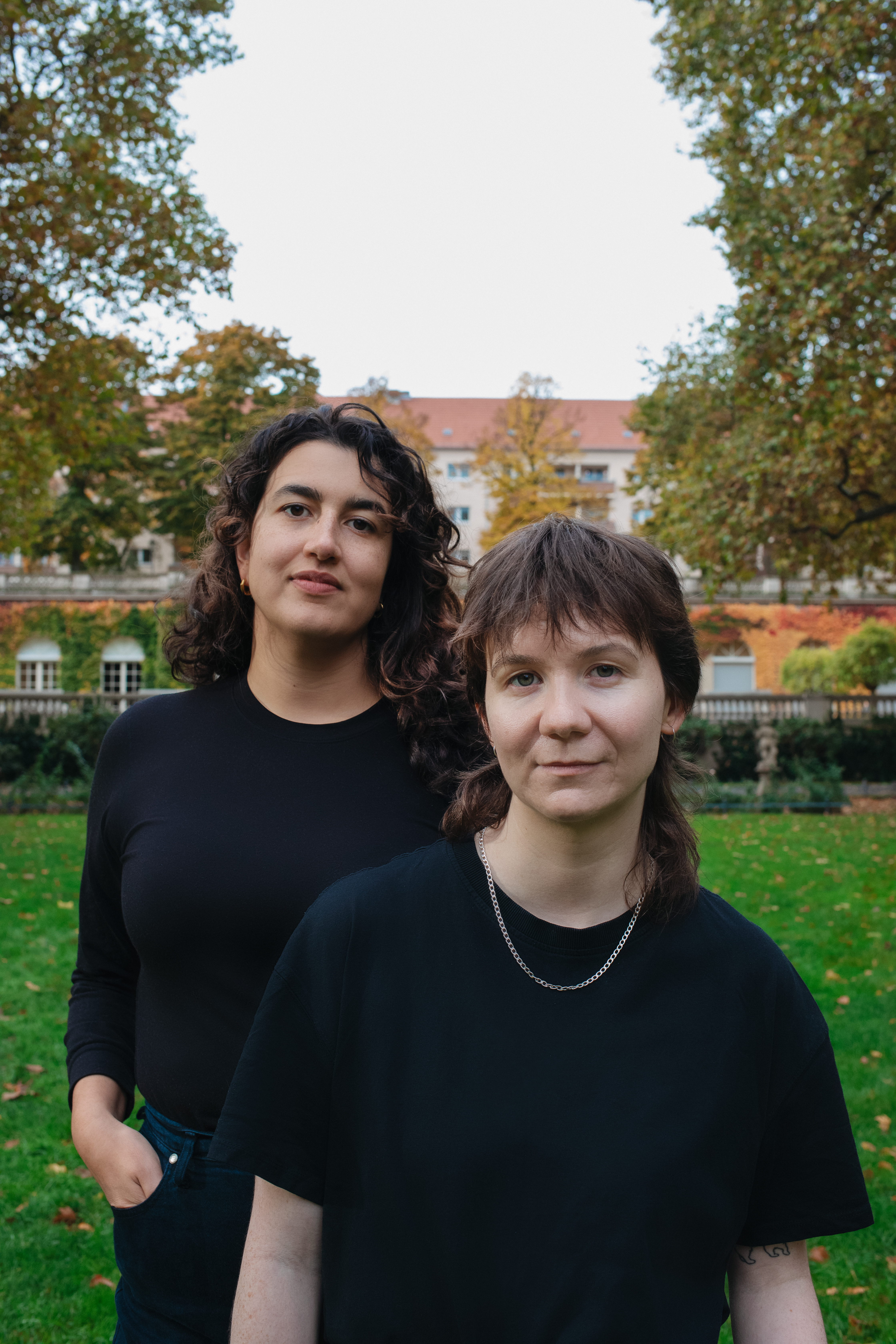 Two young persons stand in a park. Greenery from the lawn and tree leaves surrounds them, and a house is seen on the background. Both wear black and face the camera.