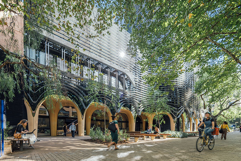 Image of students walking and cycling down brick pavement in the foreground, with the arts west building's glass facade shining in the sun in the background surrounded by leafy green trees