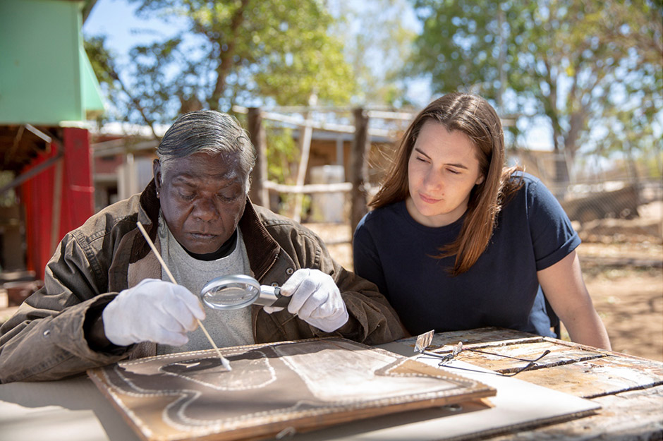 An Indigenous man and woman inspecting an art work outside in the sun with trees in the backdrop