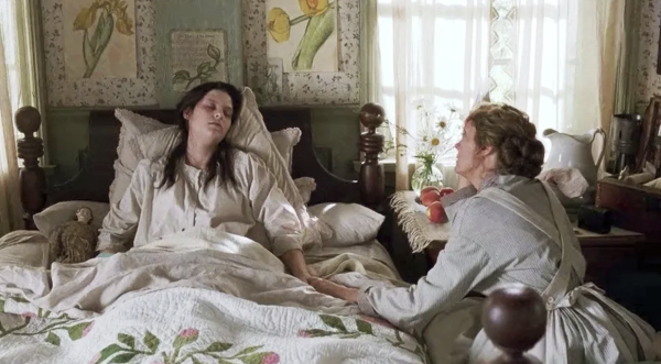 image of sickly woman with brown hair lie in bed, with floral sheets, holding hands with a caring blonde haired woman