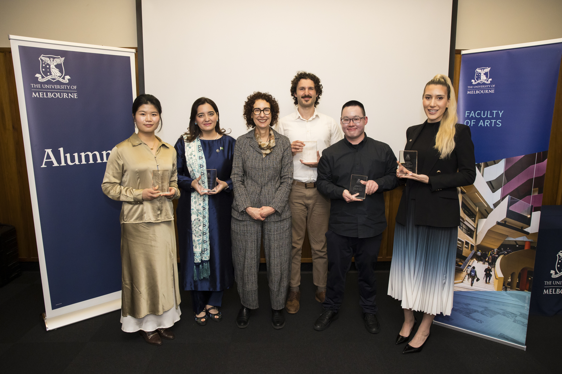 Group photo of alumni award winners standing in front of a white and timber wall, with two University of Melbourne banners each side reading alumni and Faculty of Arts respectively