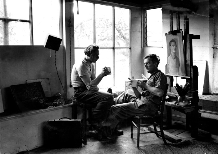 A black and white photo of two men sitting in an art studio drinking from mugs and talking