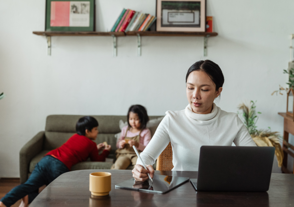 Mother working on laptop at dining table with two children playing behind her