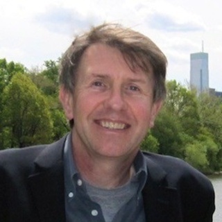 Man in blue suit jacket smiles in front of trees