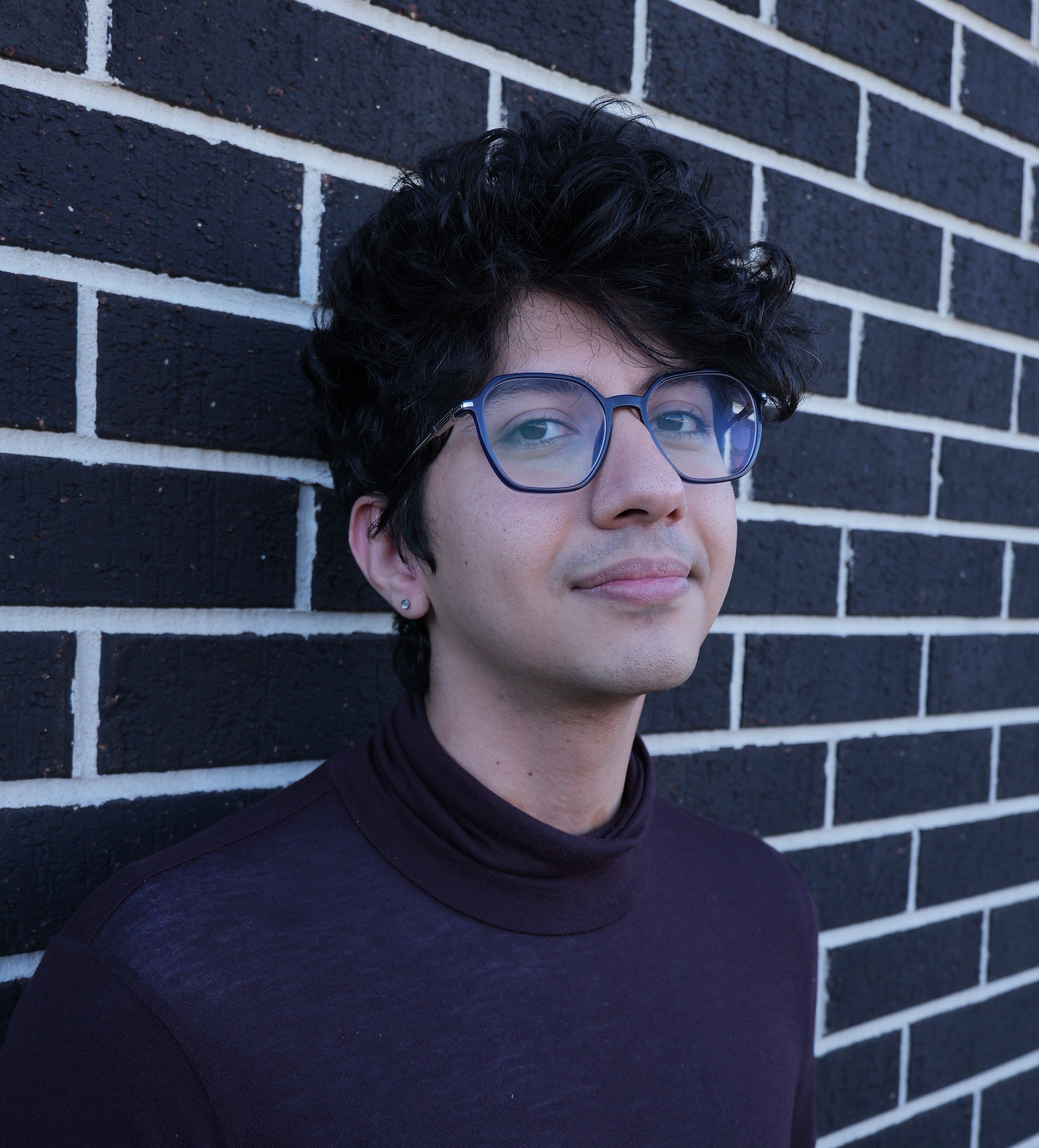 Image shows a photo of a man with dark curly hair and glasses in front of a brick wall.