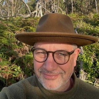 image of man wearing glasses and a brown wide brim hat in front of nature