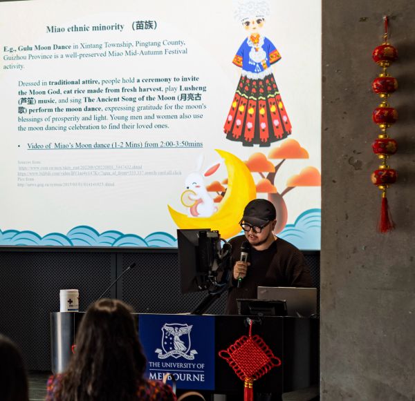 Dongyang Mi, dressed in black wearing a black cap, speaking in front of a powerpoint slide on the Miao ethnic minority in China.